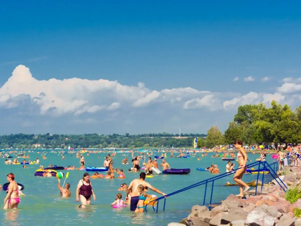Tłum ludzi cieszy się słońcem i wodą na zatłoczonej balaton plaży. Dzieci pływają na dmuchanych zabawkach, dorośli relaksują się w wodzie.
