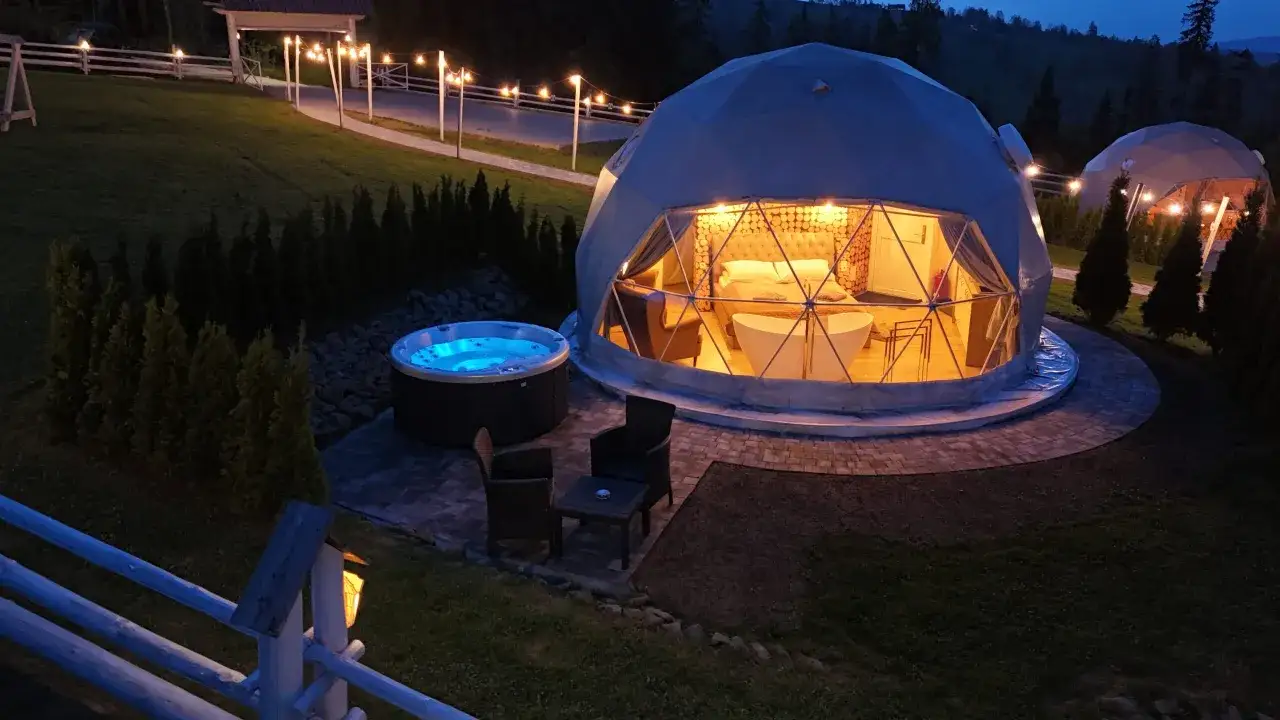 Nocny widok na luksusowe pole namiotowe w g&oacute;rach. Dwa kopułowe namioty, jeden z wanną i ł&oacute;żkiem, drugi w oddali. Obok jacuzzi i meble ogrodowe.