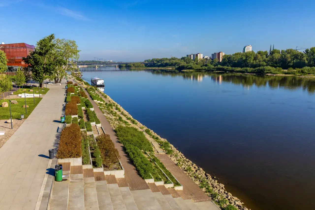 Nowoczesne miasto nad Wisłą z bulwarem, zielenią i budynkami w tle.