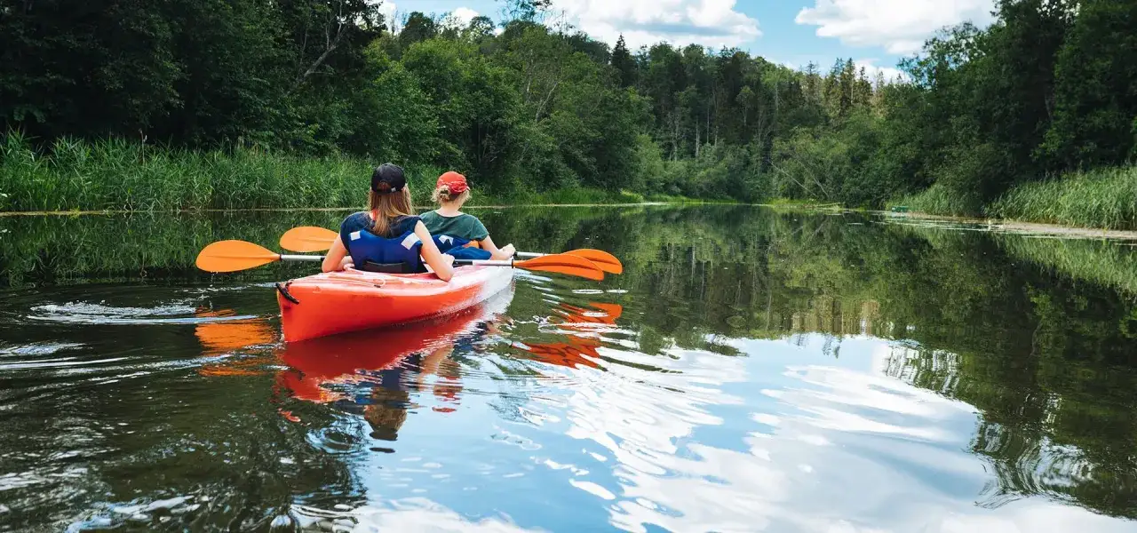 Dwie osoby w kajaku na spokojnej rzece, otoczonej zielenią. Idealne miejsce, gdzie na Mazury we dwoje, by odpocząć od zgiełku.